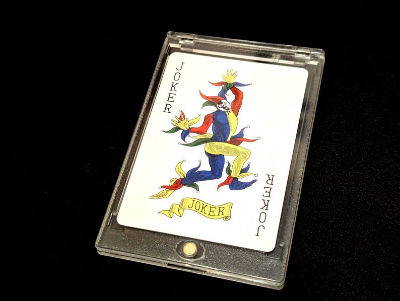May include: A Joker playing card in a clear plastic case. The card features a colorful illustration of a jester with the word "JOKER" on the top and bottom of the card and on a banner.