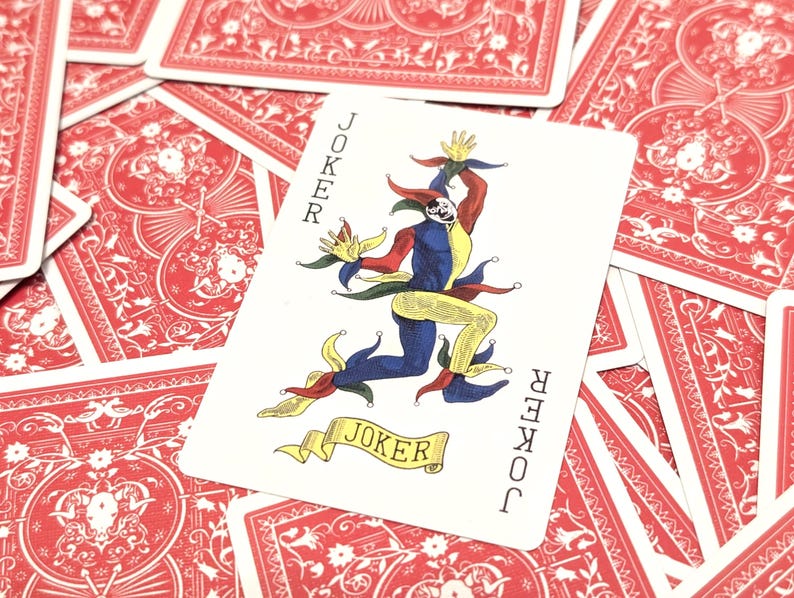 May include: A playing card featuring a colorful joker character with the word "JOKER" on the card. The card is set against a background of red playing cards with a floral design.
