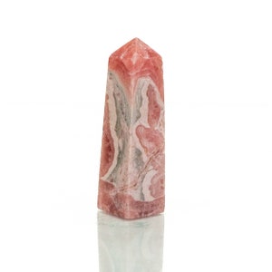 Könnte beinhalten: Ein hoher, rechteckiger Obelisk aus rosa und weißem Stein mit spitzer Spitze. Der Stein hat ein marmoriertes Aussehen mit Wirbeln aus Rosa, Weiß und Grau. Der Obelisk steht auf einer reflektierenden Oberfläche.