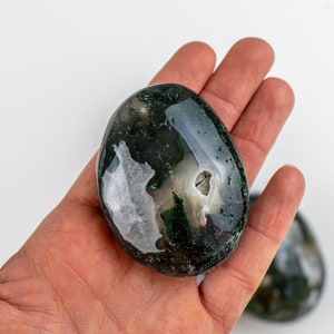 Pu&ograve; includere: Una pietra di agata muschiata lucidata, di forma ovale, tenuta in una mano. La pietra presenta un mix di motivi verde scuro e bianchi, con una qualit&agrave; traslucida. La pietra misura circa 7.6 cm di lunghezza.