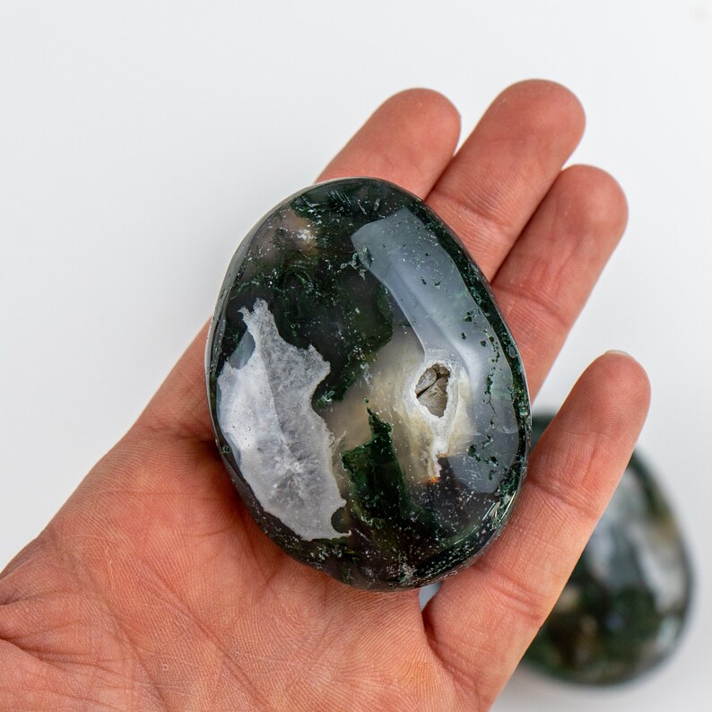 Pu&ograve; includere: Una pietra di agata muschiata lucidata, di forma ovale, tenuta in una mano. La pietra presenta un mix di motivi verde scuro e bianchi, con una qualit&agrave; traslucida. La pietra misura circa 7.6 cm di lunghezza.