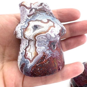 May include: A mushroom-shaped stone carving with a unique pattern of red, white, brown, and gray swirls. The mushroom is held in a hand, showcasing its detailed, natural design. The stone has a polished finish.
