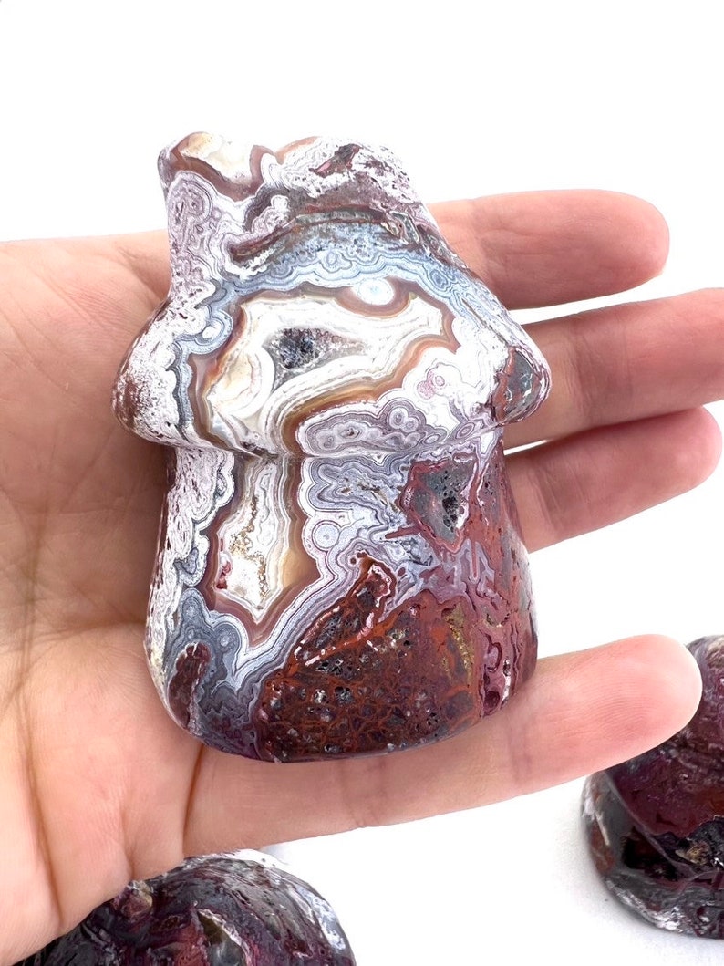 May include: A mushroom-shaped stone carving with a unique pattern of red, white, brown, and gray swirls. The mushroom is held in a hand, showcasing its detailed, natural design. The stone has a polished finish.