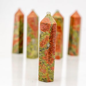 Könnte beinhalten: Eine Sammlung von Unakit-Kristall-Obelisken. Die Obelisken sind rechteckig mit einer spitzen Spitze. Der Stein hat ein gesprenkeltes Muster aus Rot, Grün und Orange. Die Obelisken stehen auf einer weißen Oberfläche.