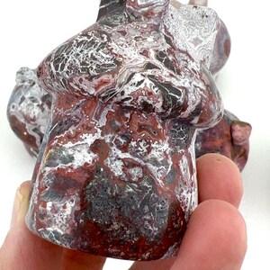 May include: A carved stone mushroom sculpture with a mottled pattern of red, white, black, and gray. The mushroom has a rounded cap and a short stem. The sculpture is held in a hand against a white background.