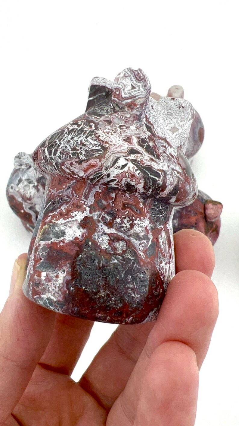 May include: A carved stone mushroom sculpture with a mottled pattern of red, white, black, and gray. The mushroom has a rounded cap and a short stem. The sculpture is held in a hand against a white background.