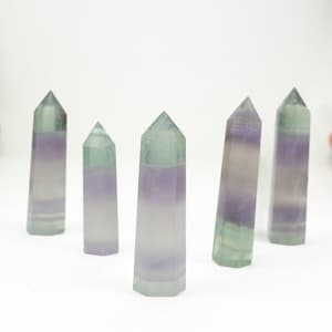 Könnte beinhalten: Fünf Fluoritturm-Kristalle. Jeder Turm hat eine sechseckige Basis. Die Kristalle sind eine Mischung aus Grün und Lila mit transluzentem Aussehen. Die Kristalle sind vor einem weißen Hintergrund angeordnet. Die Türme sind unterschiedlich hoch.