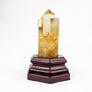 Könnte beinhalten: Ein goldgelber Citrin-Kristallpunkt, montiert auf einem dunkelrotbraunen Holzsockel. Der Kristall ist oben facettiert, und der Sockel hat ein dekoratives, mehrstufiges Design. Der Hintergrund ist schlicht weiß.