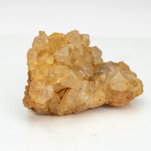 Könnte beinhalten: Eine Ansammlung von durchscheinenden, goldgelben Quarzkristallen, die auf einer rauen, beigefarbenen Mineralbasis wachsen. Das Exemplar weist zahlreiche scharfe, spitze Kristallenden auf, die das Licht einfangen und ein facettiertes Aussehen erzeugen.