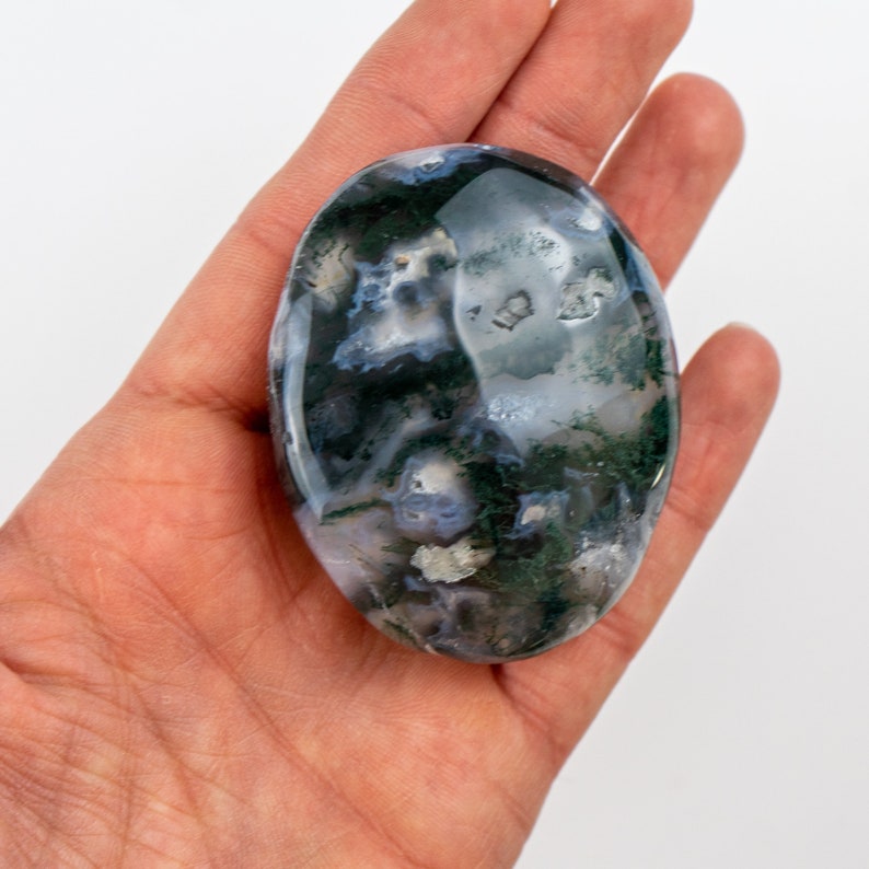 Pu&ograve; includere: Una pietra di agata muschiata lucidata, di forma ovale, con motivi bianchi traslucidi, verde scuro e blu chiaro. La pietra &egrave; tenuta in una mano su sfondo bianco, mostrando la sua bellezza naturale.
