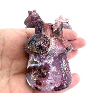 May include: A carved mushroom figurine with a textured surface. The mushroom is primarily burgundy and white with a swirling pattern. The figurine is held in a hand against a white background. Other similar figurines are visible in the background.