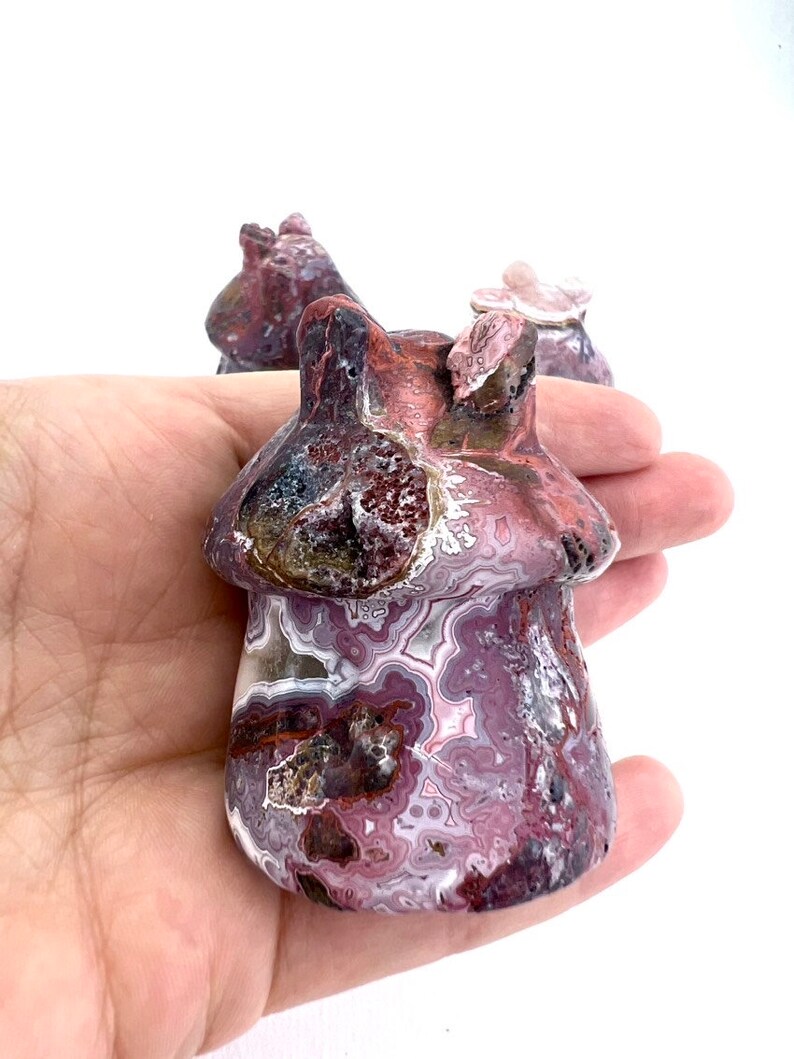 May include: A carved mushroom figurine with a textured surface. The mushroom is primarily burgundy and white with a swirling pattern. The figurine is held in a hand against a white background. Other similar figurines are visible in the background.