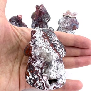 May include: A hand holds several mushroom-shaped stone carvings. The carvings are primarily red and white with intricate patterns. The front-most carving has a visible cavity. The background is white.