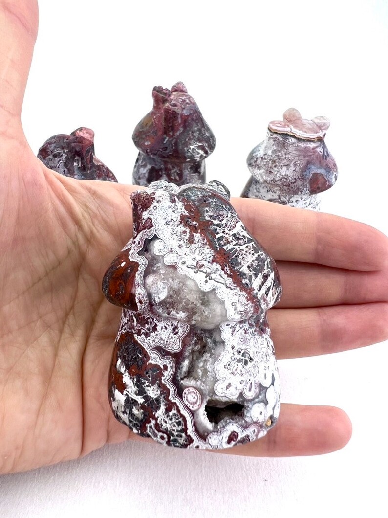 May include: A hand holds several mushroom-shaped stone carvings. The carvings are primarily red and white with intricate patterns. The front-most carving has a visible cavity. The background is white.