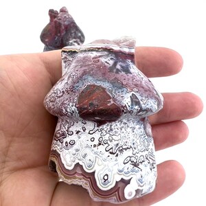May include: A mushroom-shaped stone carving with a marbled pattern of white, burgundy, and gray. The sculpture is held in a hand against a white background. A second, smaller carving is visible in the background.