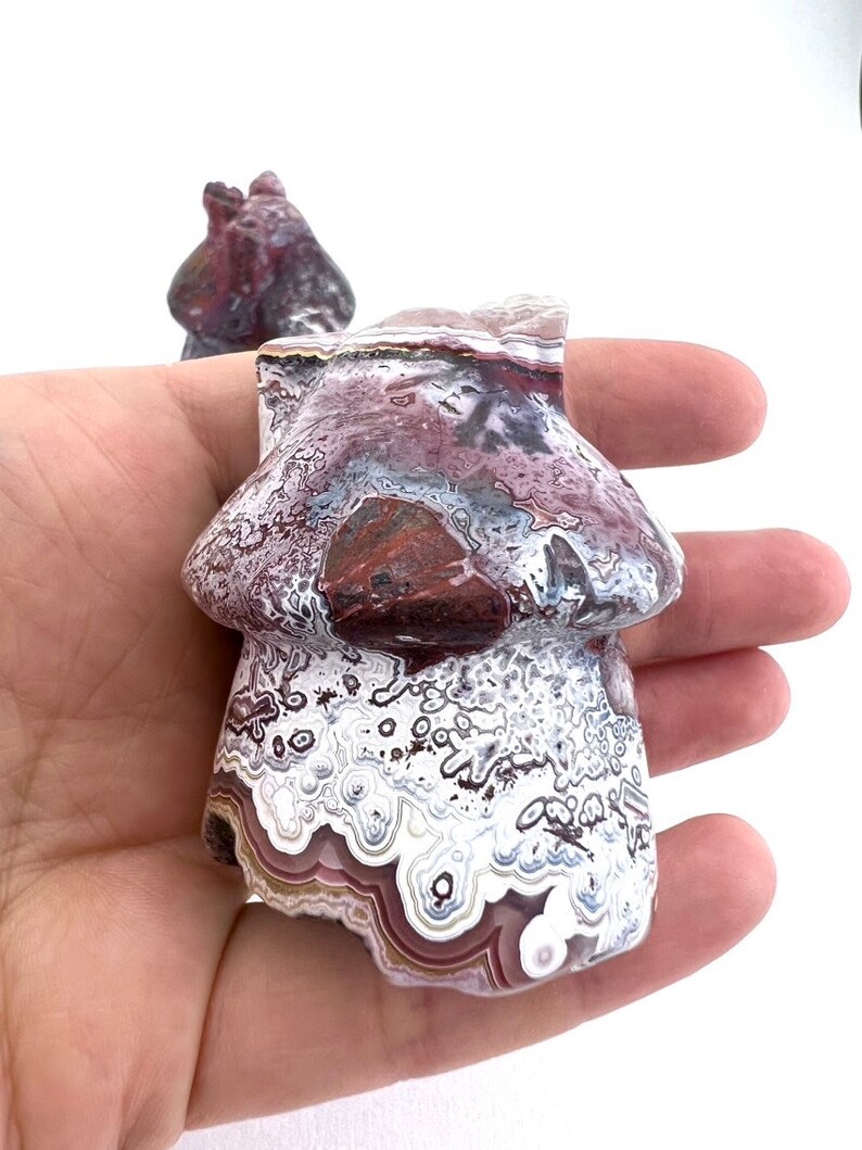 May include: A mushroom-shaped stone carving with a marbled pattern of white, burgundy, and gray. The sculpture is held in a hand against a white background. A second, smaller carving is visible in the background.