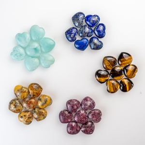 Könnte beinhalten: Eine Auswahl an herzförmigen Edelsteinen in verschiedenen Farben. Die Kollektion umfasst hellblaue, tiefblaue, braune, gelbe und violette Steine. Die Edelsteine sind in kleinen Clustern vor einem weißen Hintergrund angeordnet, um ihre einzigartigen Texturen und Farbtöne hervorzuheben.