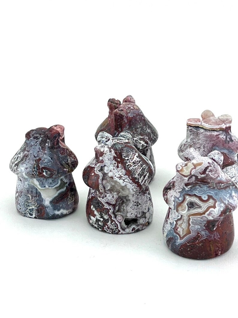 May include: A collection of five mushroom-shaped stone carvings. The carvings feature a mix of colors, including red, gray, white, and brown. The mushroom caps have a textured appearance, and the stems are smooth. The carvings are displayed on a white background.