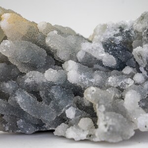 May include: Close-up of a cluster of gray and white druzy quartz crystals. The crystals have a bubbly, textured appearance, with a mix of light and dark gray hues. The background is a plain white.