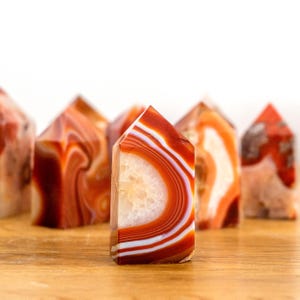 Könnte beinhalten: Eine Sammlung polierter Steintürme mit lebhaften Bändern in Orange, Rot und Weiß. Der mittlere Turm zeigt eine markante Geodenmitte mit konzentrischen Achatschichten, einem glatten, spitzen Ende und einer flachen Basis.