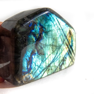 Puede incluir: Una piedra preciosa de labradorita pulida con una superficie oscura e iridiscente. La piedra muestra destellos de azul, verde y dorado. La gema se sostiene en una mano sobre un fondo blanco. Este es un artículo decorativo.