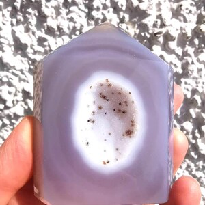 Könnte beinhalten: Ein polierter, hausförmiger Turm aus lavendelfarbenem Chalcedon mit einer zentralen, ovalen Druzy-Geodenhöhle. Das Innere der Höhle ist weiß mit kleinen, dunkelbraunen Mineraleinschlüssen, umgeben von glatten, durchscheinenden violetten Schichten.