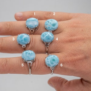 May include: Six silver rings, each featuring a large, round, light blue gemstone with white marbling. The rings are displayed on a hand, showcasing the variety of designs and stone patterns. The gemstones are the focal point.