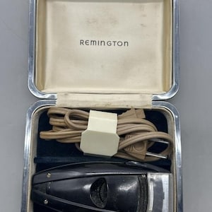 Puede incluir: Afeitadora el&eacute;ctrica Remington vintage en un estuche plateado. La afeitadora negra tiene un cabezal de l&aacute;mina plateada y un cable. El estuche est&aacute; forrado con terciopelo azul y tiene el nombre de la marca "Remington" impreso en el interior.