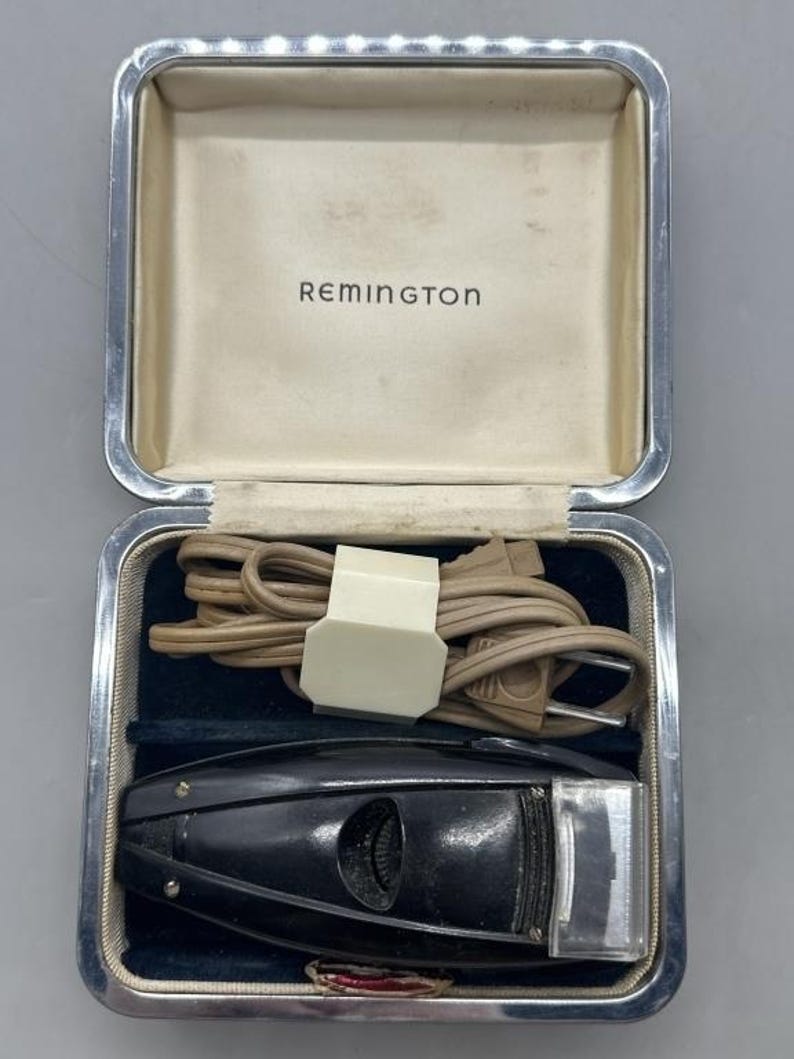 Puede incluir: Afeitadora el&eacute;ctrica Remington vintage en un estuche plateado. La afeitadora negra tiene un cabezal de l&aacute;mina plateada y un cable. El estuche est&aacute; forrado con terciopelo azul y tiene el nombre de la marca "Remington" impreso en el interior.