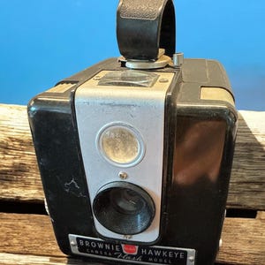 May include: A black and silver Brownie Hawkeye Flash camera with a black leather strap. The camera has a red circle with the word "Flash" on it.