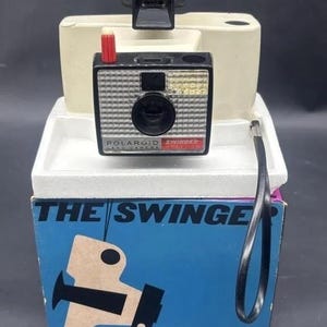 May include: A vintage Polaroid Swinger instant camera, black and silver with a red button, atop a white base. The camera is on a blue box with the words "THE SWINGER" and a graphic design.