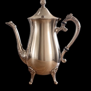 May include: A polished silver-toned coffee pot featuring a tall, tapered body, a curved spout with ornate detailing, and a matching lid with a finial. The vessel stands on three decorative feet and includes a handle with black accents.