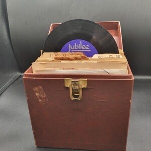 May include: A brown wooden record storage box with a metal clasp, filled with vinyl records. One record is visible, with a blue label that reads "Jubilee". The box contains several record sleeves with numbers.