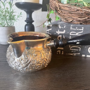 May include: A silver-colored, ornate coffee pot with a floral design. The pot has a spout and a black wooden handle. The pot is sitting on a dark wooden surface. The pot is likely used for brewing coffee or tea.