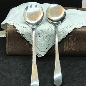 May include: Two silver serving spoons. One spoon has a rounded bowl with three tines, and the other has a rounded bowl. Both spoons have long, slender handles. They rest on a white lace-edged cloth, with a dark brown book in the background.
