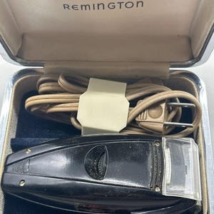 Puede incluir: Afeitadora el&eacute;ctrica Remington vintage en un estuche plateado. La afeitadora negra tiene una cubierta de cabeza de pl&aacute;stico transparente y un dial. El estuche est&aacute; abierto, revelando un cable de alimentaci&oacute;n beige enrollado y el logotipo de Remington.