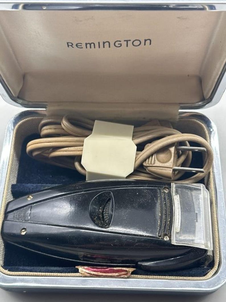 Puede incluir: Afeitadora el&eacute;ctrica Remington vintage en un estuche plateado. La afeitadora negra tiene una cubierta de cabeza de pl&aacute;stico transparente y un dial. El estuche est&aacute; abierto, revelando un cable de alimentaci&oacute;n beige enrollado y el logotipo de Remington.