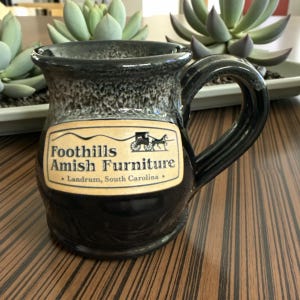 May include: A black and gray ceramic mug with a handle. The mug features a label with the text "Foothills Amish Furniture" and an illustration of a horse-drawn carriage. The mug is sitting on a wooden surface.