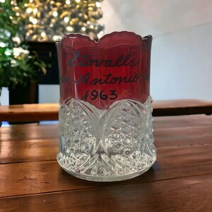 May include: A clear glass cup with a red glass insert. The red glass insert has the text "Stornall's San Antonio 1963" written in black.