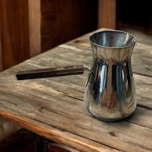 May include: A silver coffee pot with a wooden handle sits on a rustic wooden surface.