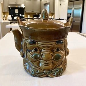 May include: A brown ceramic teapot with a whimsical face design. The teapot features a smiling face with round glasses, a bow tie, and a lid with a handle. The teapot is a unique and decorative kitchen item.