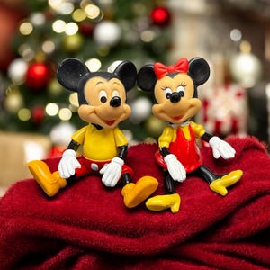 Pode incluir: Duas figuras de Mickey e Minnie Mouse sentadas numa superf&iacute;cie vermelha. Mickey usa camisa amarela, cal&ccedil;&otilde;es vermelhos e sapatos amarelos. Minnie usa um vestido vermelho, sapatos amarelos e um la&ccedil;o vermelho. Uma &aacute;rvore de Natal ao fundo.