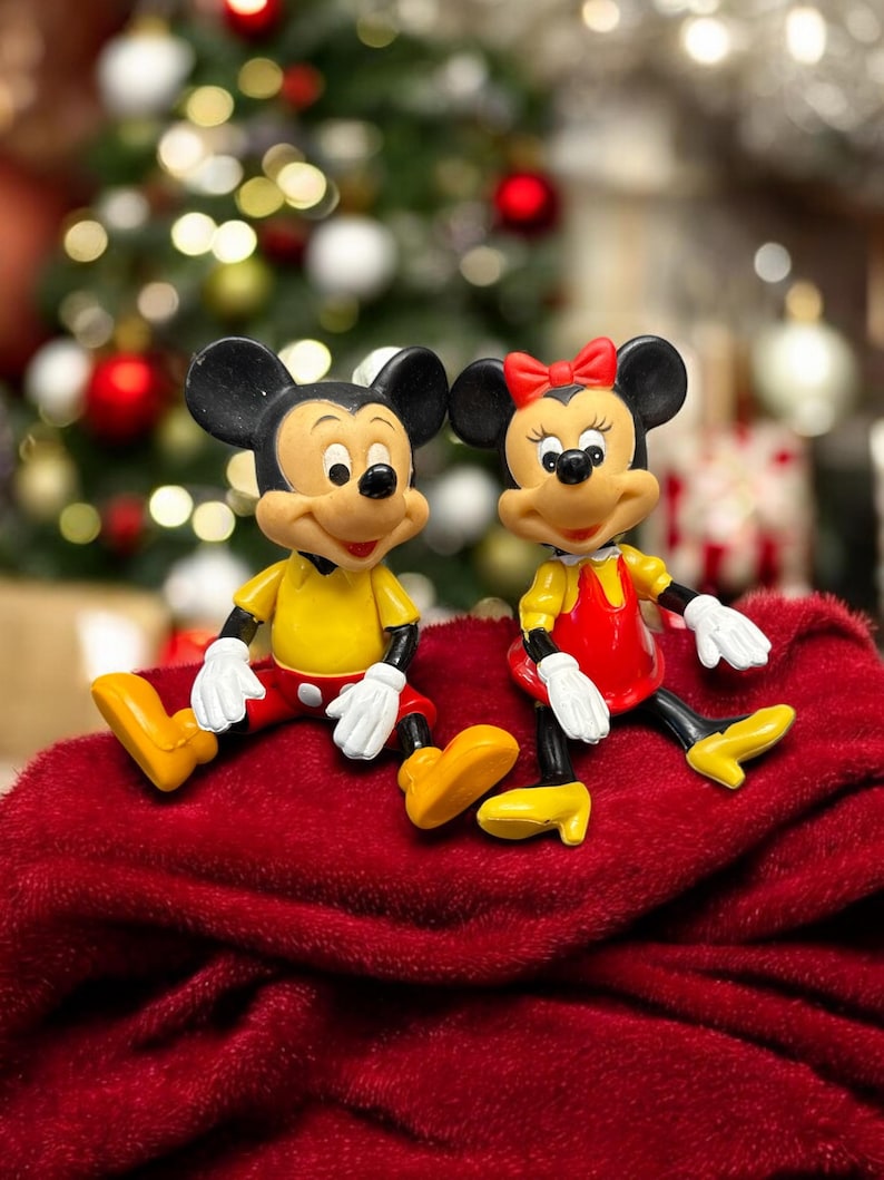 Pode incluir: Duas figuras de Mickey e Minnie Mouse sentadas numa superf&iacute;cie vermelha. Mickey usa camisa amarela, cal&ccedil;&otilde;es vermelhos e sapatos amarelos. Minnie usa um vestido vermelho, sapatos amarelos e um la&ccedil;o vermelho. Uma &aacute;rvore de Natal ao fundo.