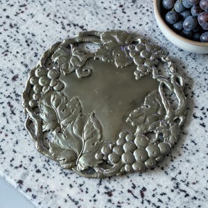 May include: A silver-colored metal trivet with a grape and leaf design. The trivet is round with a central flat area and a detailed border featuring clusters of grapes and grape leaves. The trivet is on a speckled gray surface.