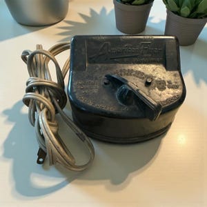May include: A black American Flyer transformer with a dial and attached power cord. The transformer has text that reads "American Flyer" and "The A.C. Gilbert Co." The cord is beige.