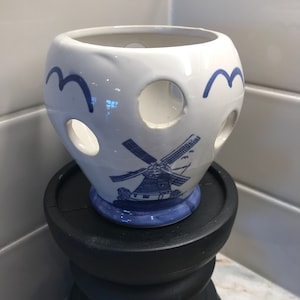 May include: A white ceramic vase with blue and white windmill design. The vase has holes around the sides and is sitting on a black wooden pedestal.