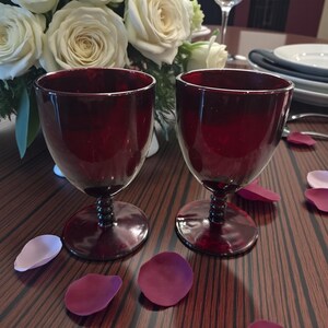 May include: Two ruby red wine glasses with a bulbous bowl and a decorative stem sit on a wooden table. Rose petals in shades of pink and burgundy are scattered around the glasses. A bouquet of white roses is in the background.