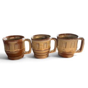 May include: Three ceramic mugs arranged in a row, each featuring a cylindrical body with a vertical panel design, a side handle, and a two-tone brown and tan glazed finish that transitions from a darker rim to a lighter base.