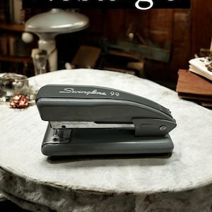 May include: A gray Swingline 99 stapler sits on a round table covered with a white cloth. The word "Nostalgia" is displayed in a black rectangle above the stapler. The stapler is a vintage office supply.