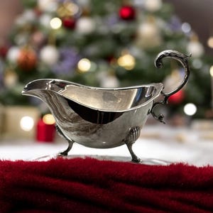 May include: A silver gravy boat with a decorative handle and three legs. The boat is sitting on a red fuzzy surface.